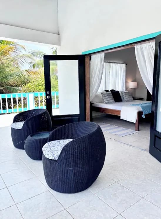Woven armchairs on a tiled veranda, its doors open onto a bedroom with a curtained four-poster bed