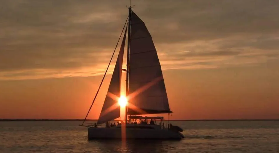 A catamaran under full sail, silhouetted against an orange sunset over the water