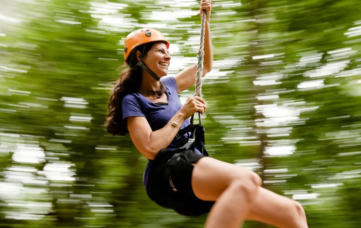 A smiling woman in a helmet flying along a zip line through the jungle