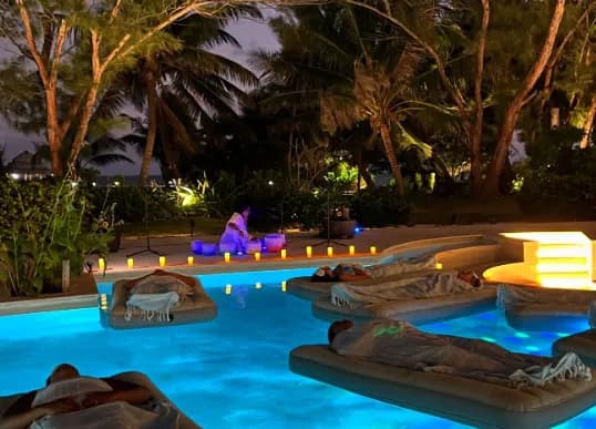 Guests lying on floating mats in a pool lit blue at dusk, surrounded by candles and palms