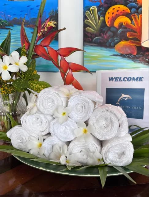 Rolled white towels on a tray of palm leaves and frangipani, beside a framed welcome card