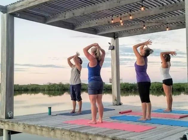 Four women holding a yoga pose on a covered deck over the water at dusk